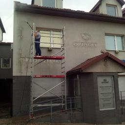 Błysk Paweł Szczepaniak - Pracownik na rusztowaniu myje elewację budynku firmy Optomet za pomocą myjki ciśnieniowej, widoczne ślady czyszczenia na ścianie.