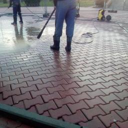 Błysk Paweł Szczepaniak - Czyszczenie kostki brukowej myjką ciśnieniową, widoczne dwie osoby w roboczych ubraniach i gumowych butach, w tle żółta myjka ciśnieniowa.