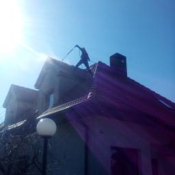 Błysk Paweł Szczepaniak - Pracownik w kombinezonie myje dach domu myjką ciśnieniową na tle błękitnego nieba i silnego słońca, widoczna lampa uliczna i fragment elewacji.