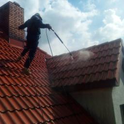 Błysk Paweł Szczepaniak - Pracownik w kombinezonie i kasku czyści czerwoną dachówkę za pomocą myjki ciśnieniowej na tle błękitnego nieba z chmurami.