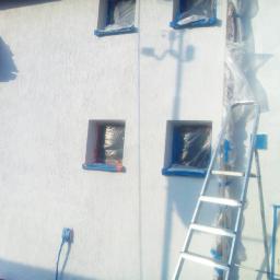 Błysk Paweł Szczepaniak - Biała elewacja budynku z zabezpieczonymi folią i taśmą okiennicami, przygotowana do prac wykończeniowych, z widoczną aluminiową drabiną opartą o ścianę.