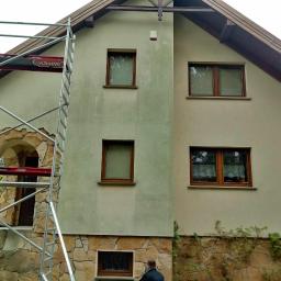 Błysk Paweł Szczepaniak - Dwukondygnacyjny dom z elewacją w trakcie czyszczenia, widoczne różnice w kolorze na ścianie, rusztowanie Altrex przy wejściu z kamienną obudową, fragment osoby w dolnej części kadru.