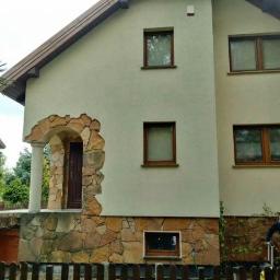 Błysk Paweł Szczepaniak - Dwupiętrowy dom z beżową elewacją i kamiennym wykończeniem wokół drzwi wejściowych i na fundamencie, z rusztowaniem po prawej stronie i mężczyzną w kurtce obsługującym myjkę ciśnieniową.