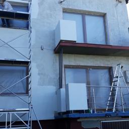Błysk Paweł Szczepaniak - Pracownik na rusztowaniu maluje elewację domu jednorodzinnego na biało, okna zabezpieczone folią, widoczny fragment ogrodu z workiem zaprawy.