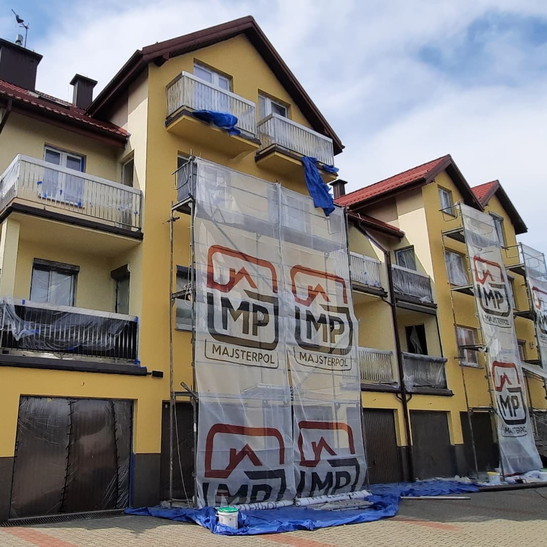 Żółty budynek wielorodzinny w trakcie prac elewacyjnych, osłonięty siatkami z logo firmy. Widoczne rusztowania, zabezpieczenia okien folią i plandeka na ziemi. Część budynku jest już pomalowana.