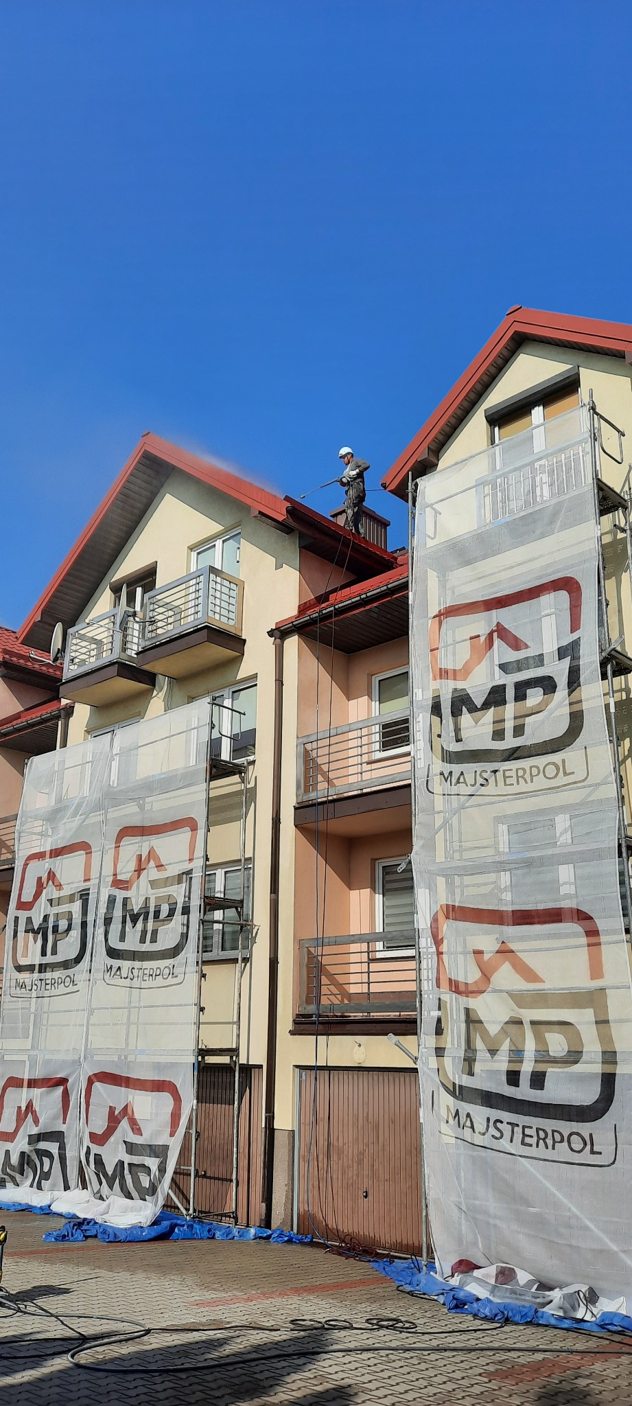Pracownik w kombinezonie i kasku czyści dach budynku wielorodzinnego za pomocą myjki ciśnieniowej; na elewacji widoczne rusztowania z banerami reklamowymi firmy Majsterpol.