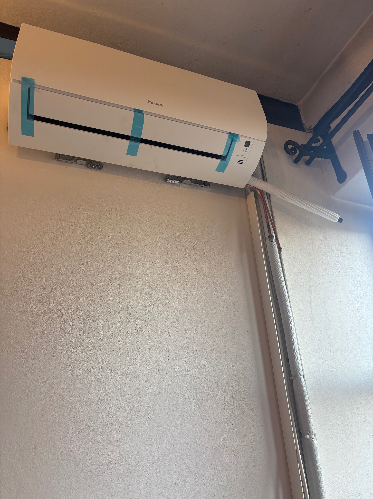 Zamontowany klimatyzator Daikin z niebieską taśmą zabezpieczającą, widoczne rury instalacyjne w białej osłonie, na jasnej ścianie.