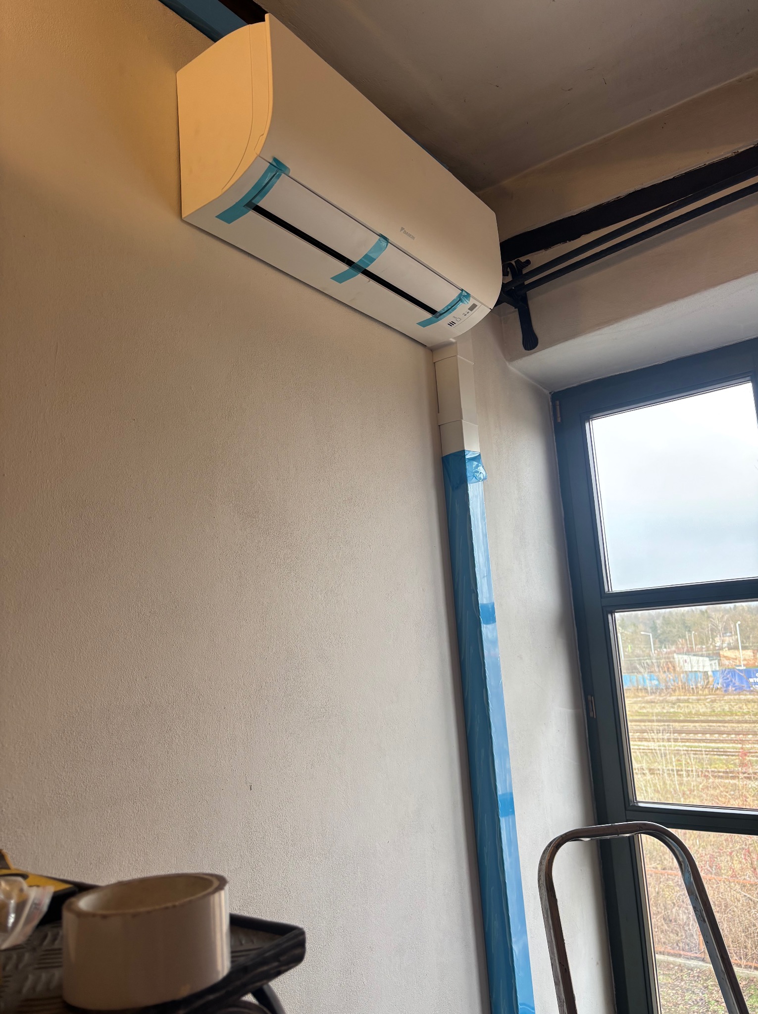 Zamontowany klimatyzator Daikin na ścianie, zabezpieczony niebieską folią ochronną, z widoczną instalacją i oknem w tle. Widoczny fragment drabiny.