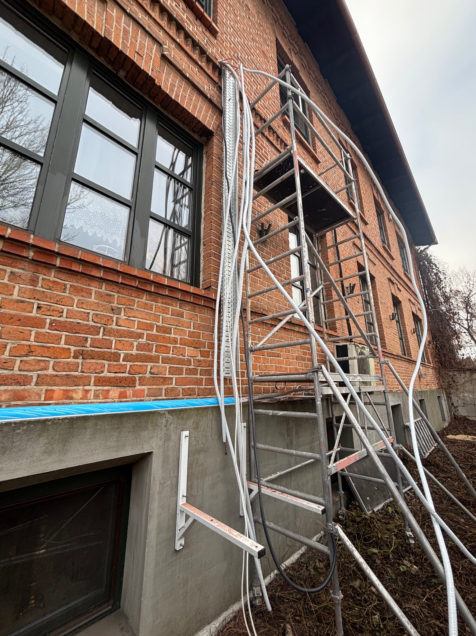 Ceglana elewacja budynku w trakcie prac, widoczne rusztowanie i rury. Ochrona okien folią. Ujęcie z dołu, pochmurne niebo.