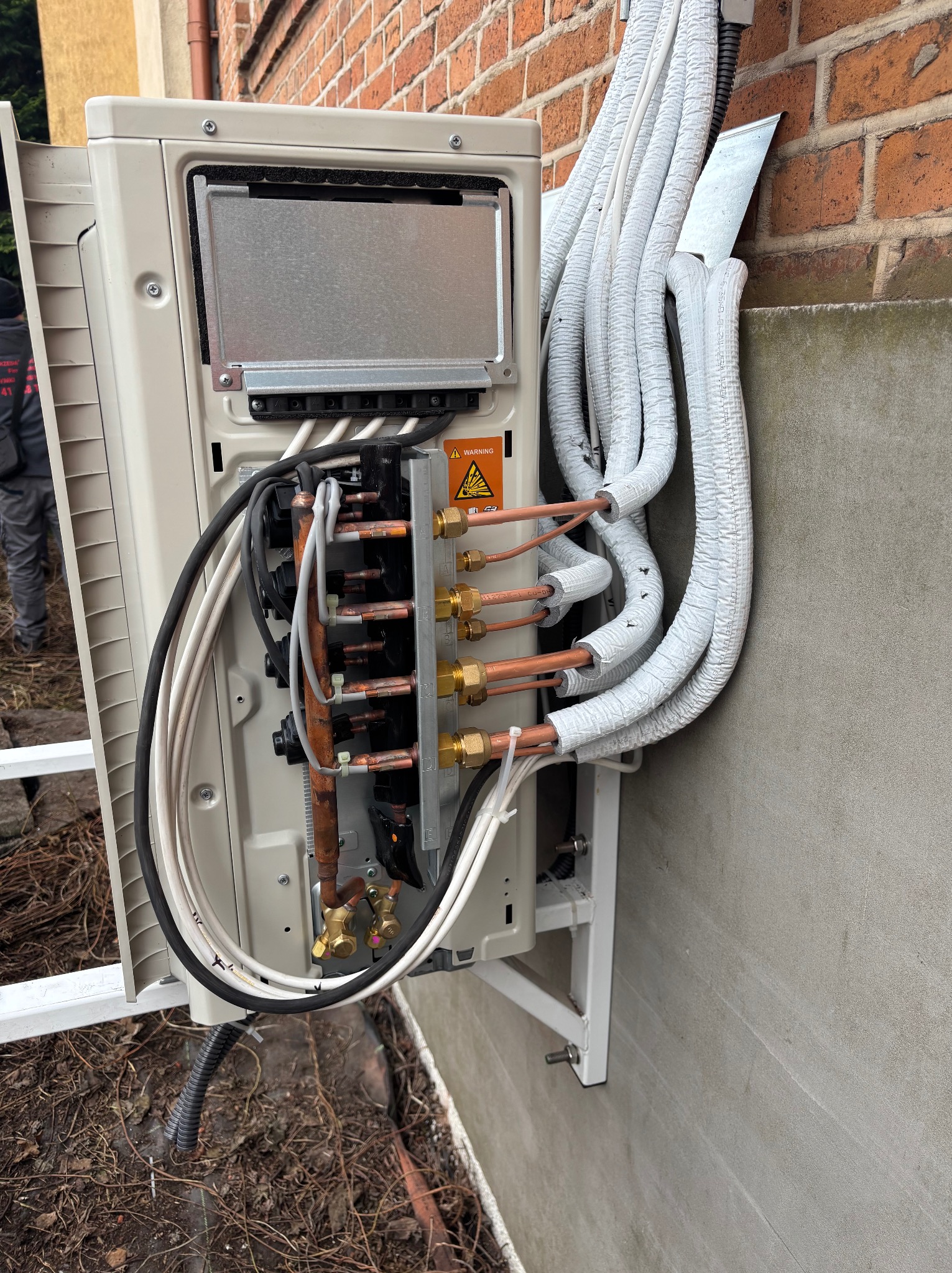 Zamontowana zewnętrzna jednostka klimatyzacji z widocznymi rurami miedzianymi i przewodami elektrycznymi, przymocowana do ściany budynku.