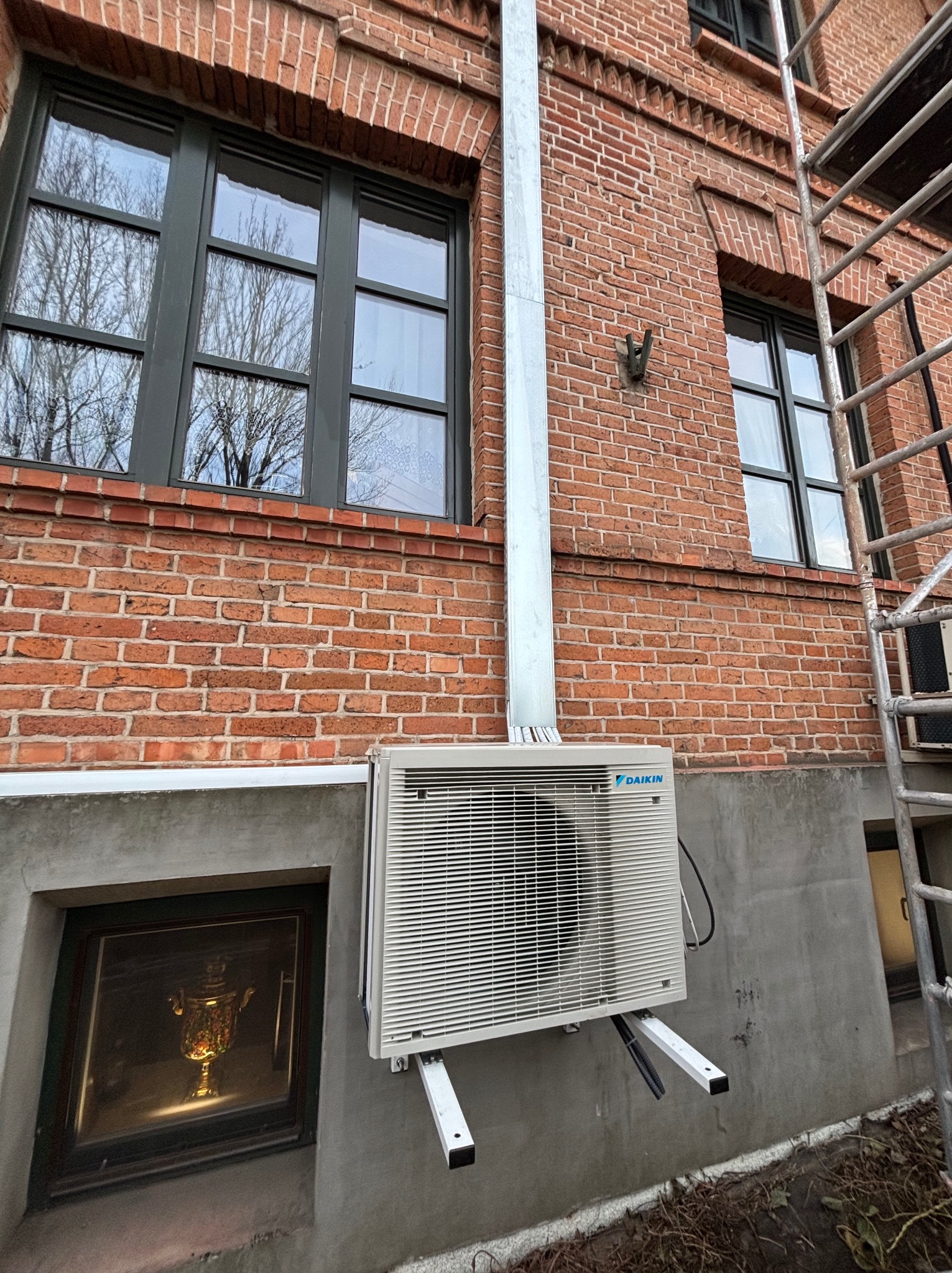 Zamontowany klimatyzator Daikin na ceglanej ścianie budynku z widoczną rurą odprowadzającą i fragmentem rusztowania. W tle okno z odbiciem drzew.