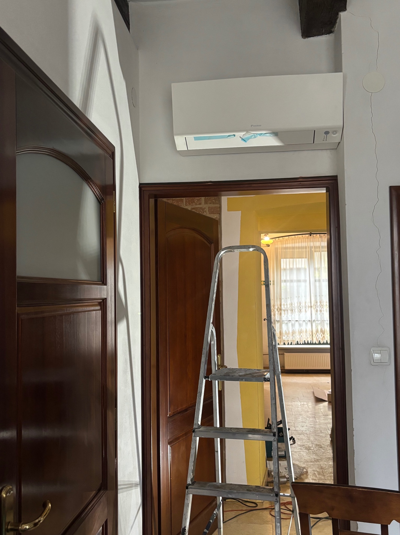 Wnętrze w trakcie malowania: drabina oparta o ścianę z żółtymi pasami, widoczny klimatyzator i drewniane drzwi. Prace malarskie w toku, widok na okno z firankami.
