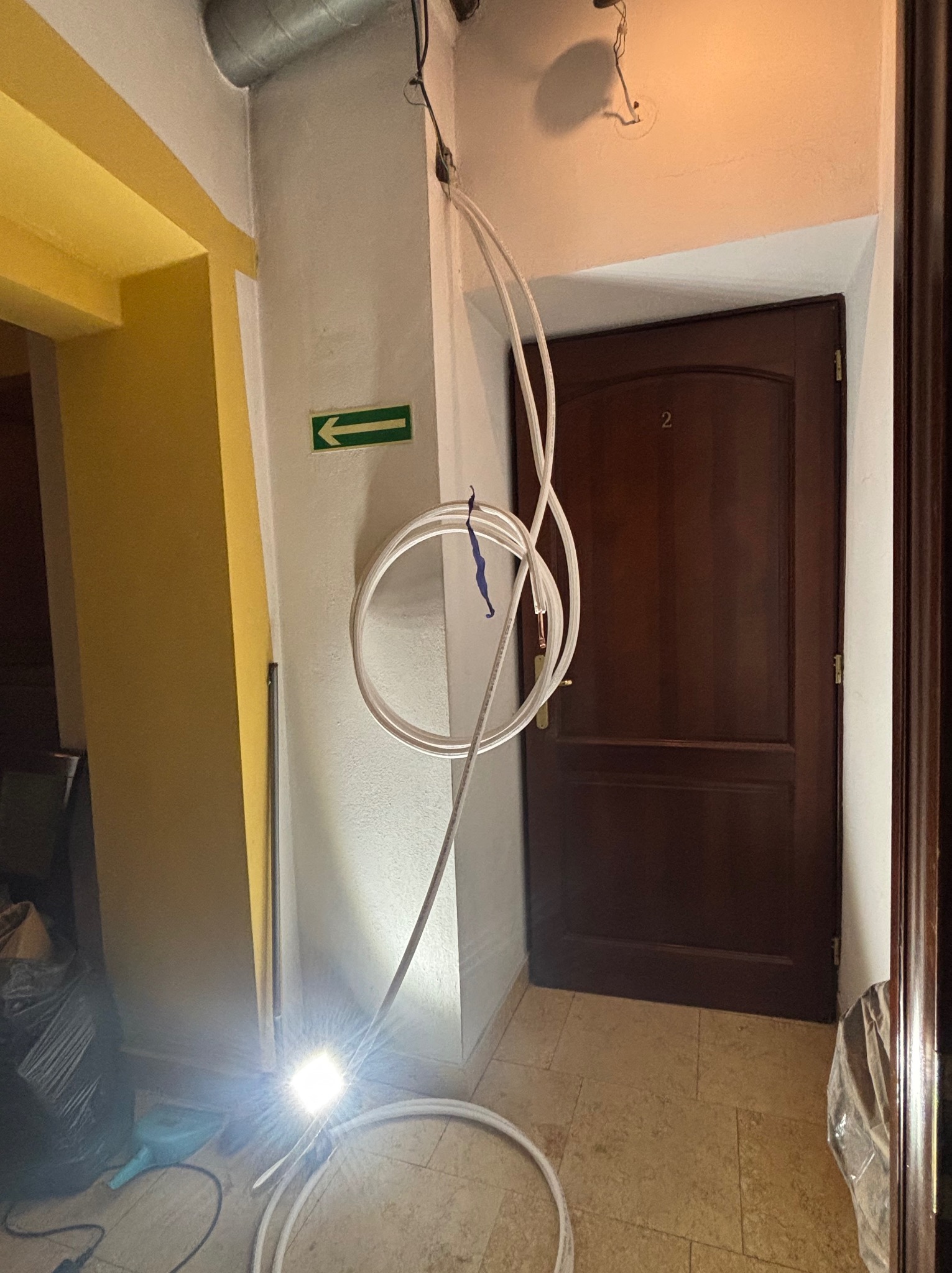 Białe rury zwisają ze ściany w korytarzu, obok brązowych drzwi z numerem 2. Widoczny fragment żółtej ściany i podłoga z płytek. Strzałka kierunkowa na ścianie.