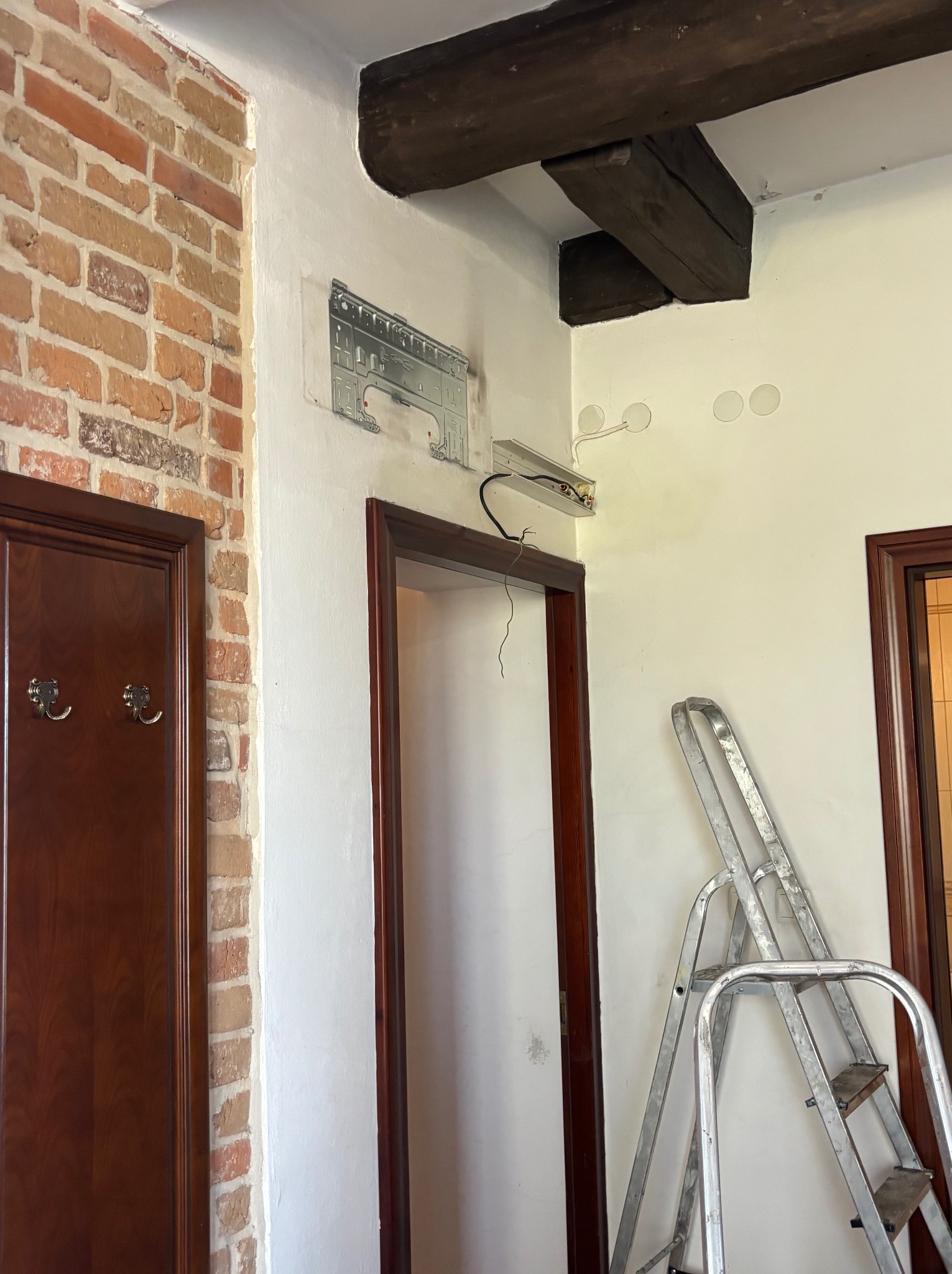 Wnętrze w trakcie remontu: widoczna ściana z cegły, przygotowane miejsce na klimatyzator, otwory na oświetlenie i drabina oparta o ścianę. Drewniane belki stropowe.