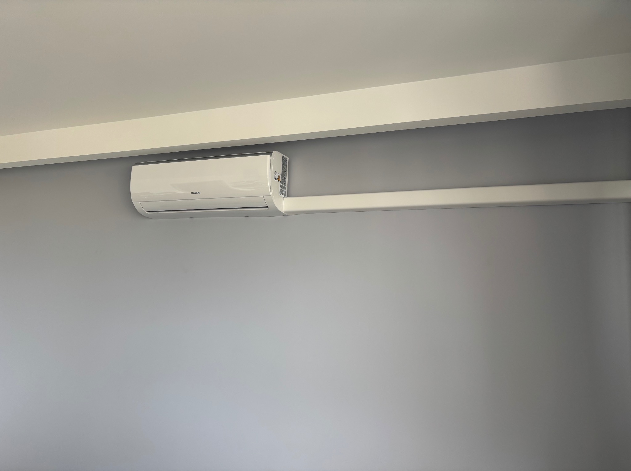 Biały klimatyzator marki Kaisai zamontowany na szarej ścianie, z widoczną białą listwą przypodłogową i fragmentem sufitu. Minimalistyczny design wnętrza.