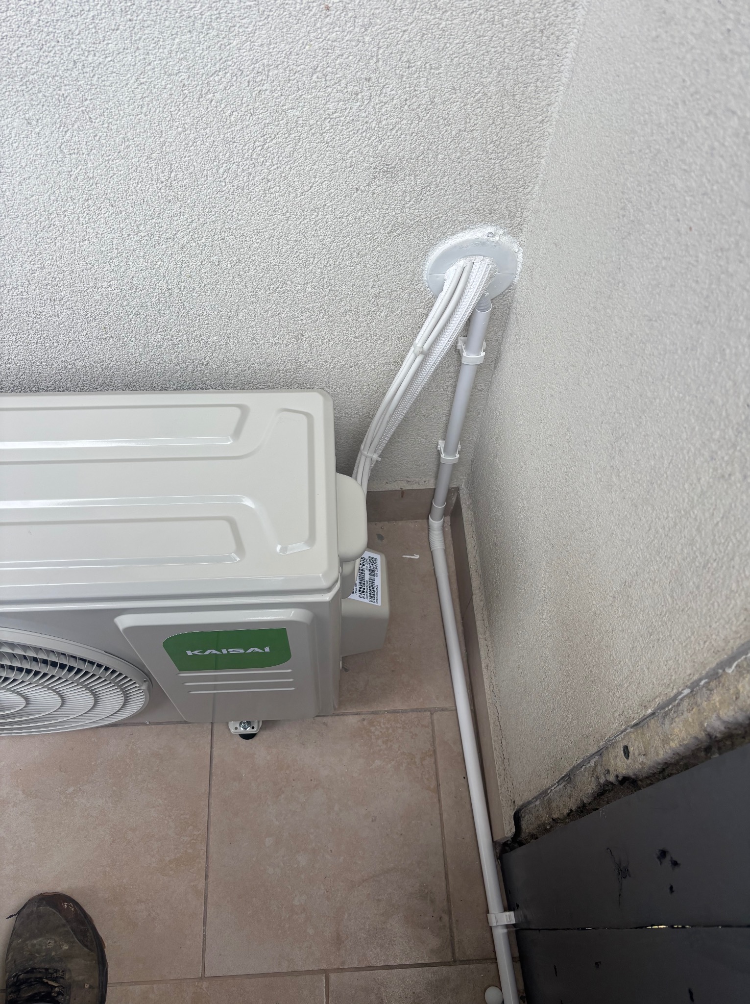 Klimatyzator Kaisai zamontowany na zewnątrz budynku, z widocznymi przewodami i rurami odprowadzającymi, przymocowanymi do ściany. Widoczny fragment buta w dolnym rogu.