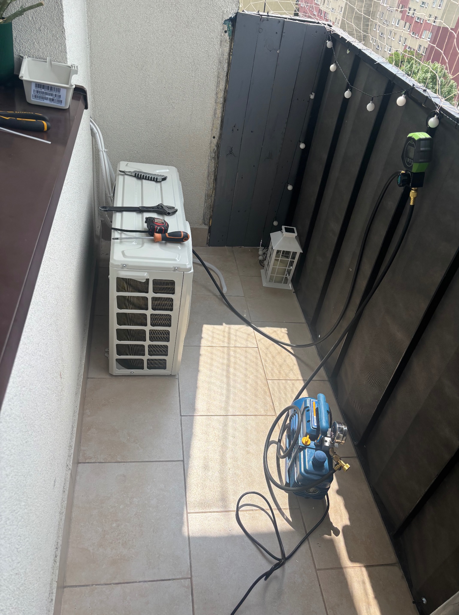 Kompaktowy klimatyzator na balkonie z narzędziami i pompą próżniową, gotowy do montażu. Widoczne oświetlenie dekoracyjne i elementy wykończenia balkonu.