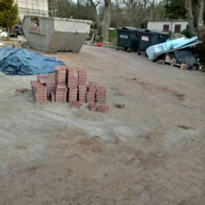 Stosy czerwonej kostki brukowej ułożone na ziemi, w tle kontener na śmieci i inne odpady budowlane.