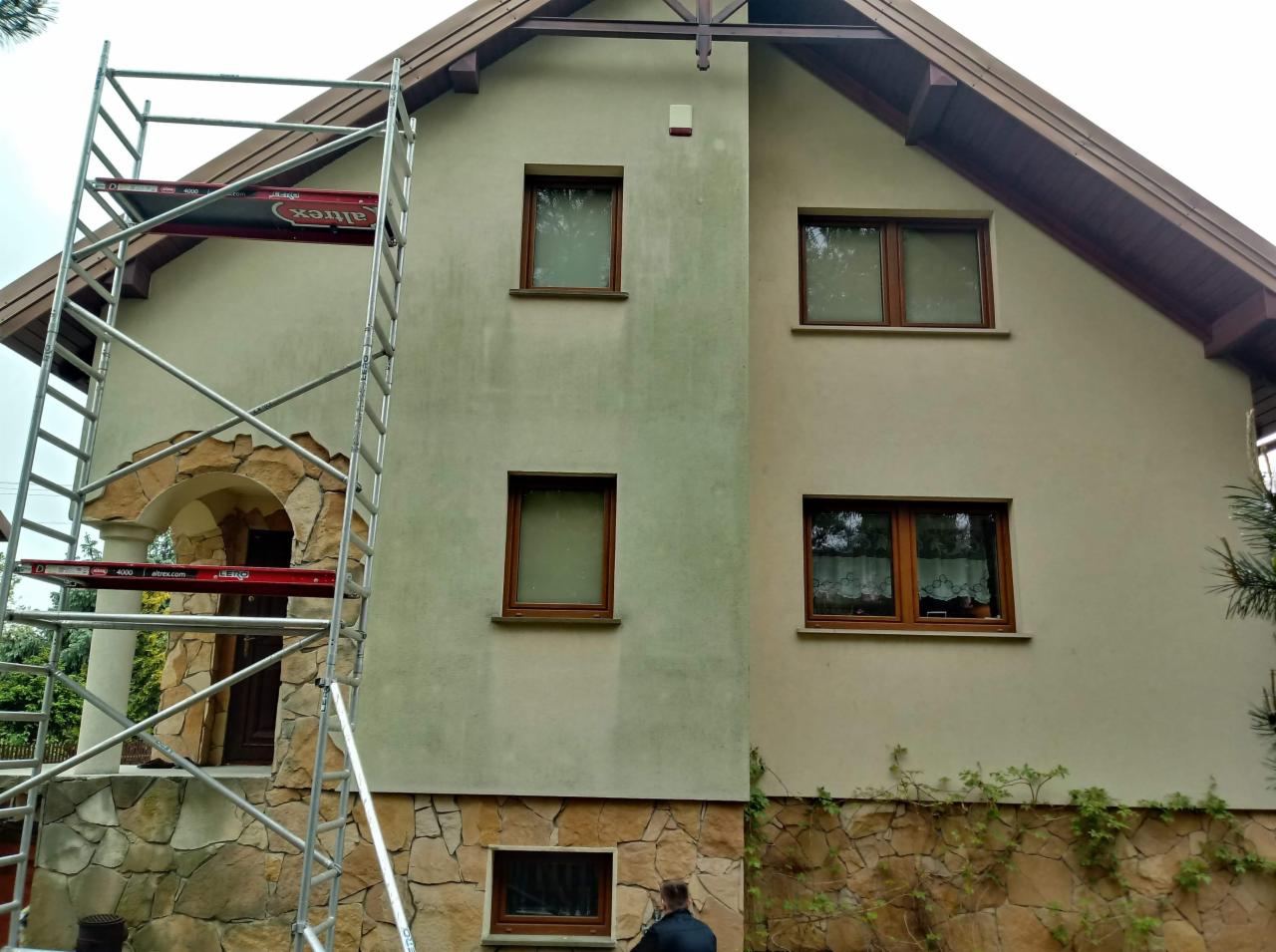 Dwukondygnacyjny dom z elewacją w trakcie czyszczenia, widoczne różnice w kolorze na ścianie, rusztowanie Altrex przy wejściu z kamienną obudową, fragment osoby w dolnej części kadru.