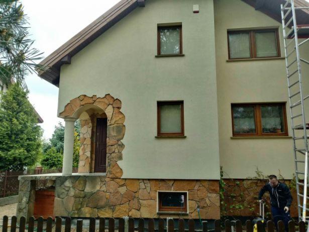 Dwupiętrowy dom z beżową elewacją i kamiennym wykończeniem wokół drzwi wejściowych i na fundamencie, z rusztowaniem po prawej stronie i mężczyzną w kurtce obsługującym myjkę ciśnieniową.