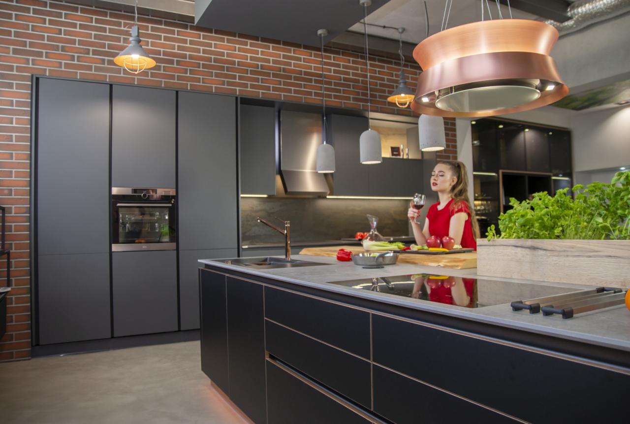 Nowoczesna kuchnia w szarościach z ceglaną ścianą, wyspą kuchenną z blatem z szarego kamienia i miedzianymi akcentami, oświetlona wiszącymi lampami. Kobieta w czerwonej bluzce trzyma kieliszek wina.