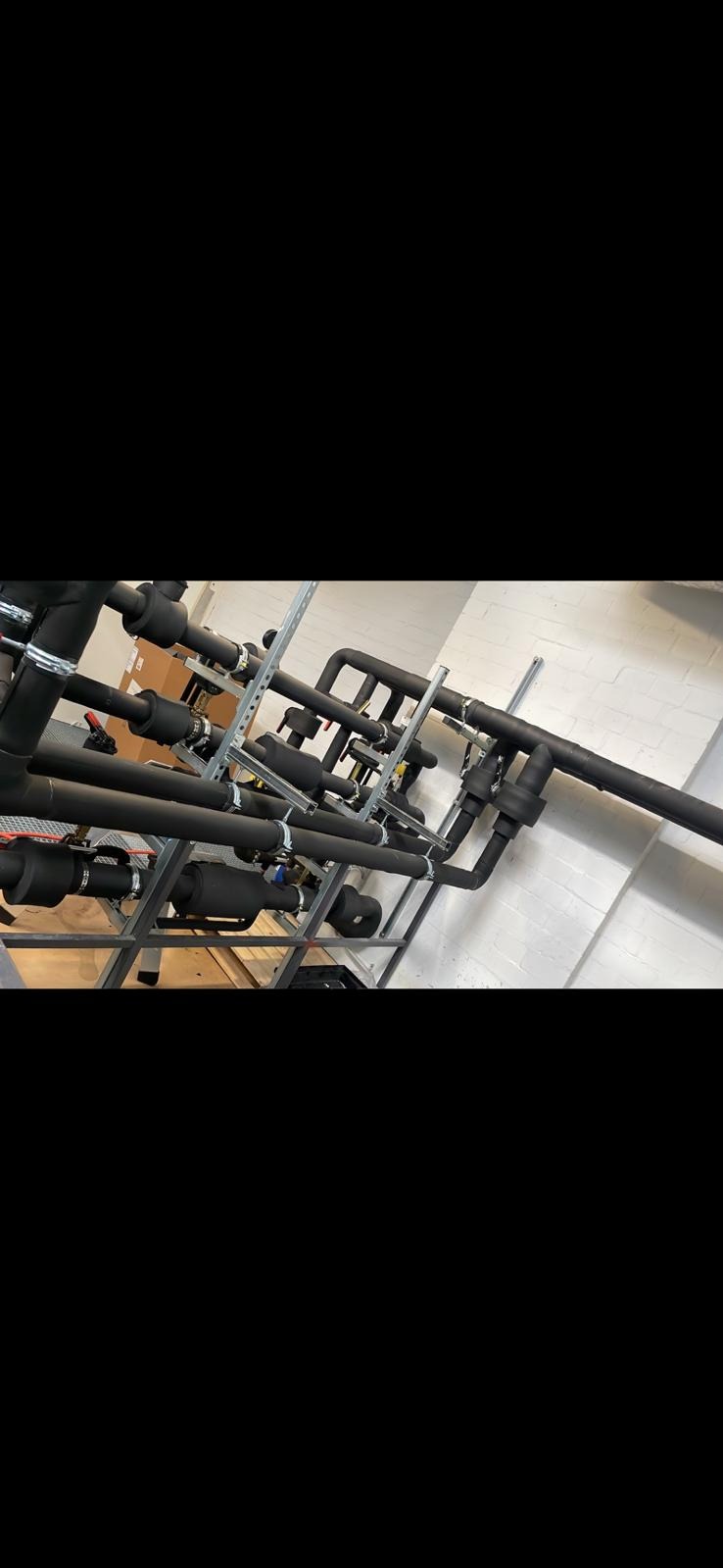 Izolowane rury instalacji grzewczej, zamocowane na metalowym stelażu przy białej ścianie. Widoczne zawory i elementy łączeniowe. Czarna izolacja termiczna.