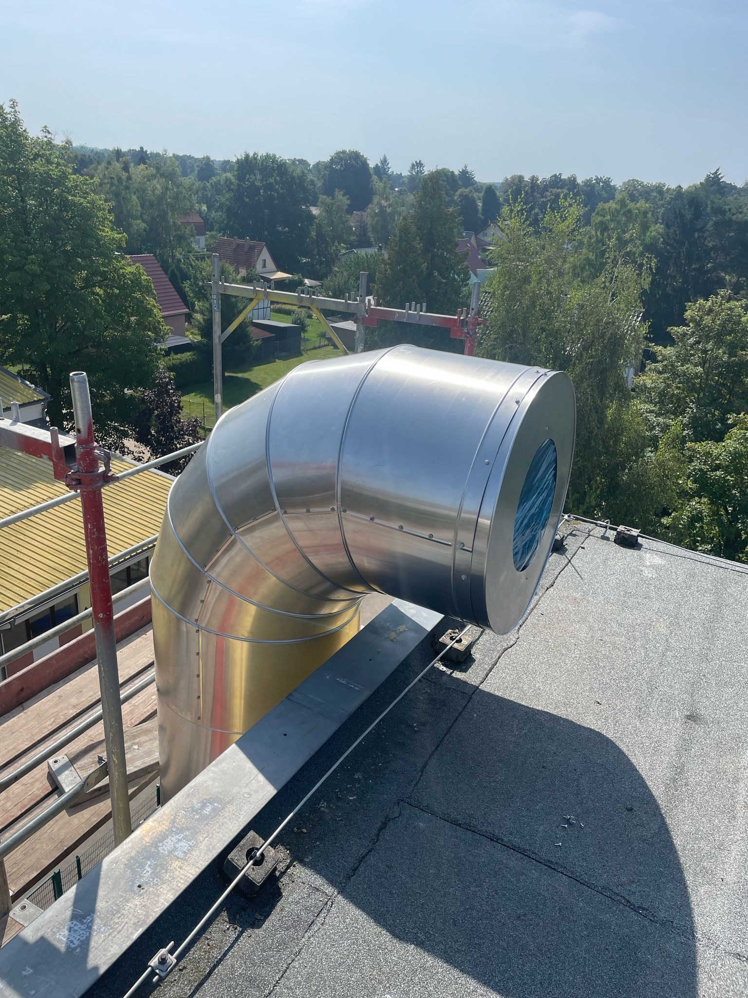 Błyszczący, metalowy komin wentylacyjny na dachu domu z widocznym zabezpieczeniem wewnątrz, na tle zieleni drzew i błękitnego nieba. Widoczne rusztowanie.
