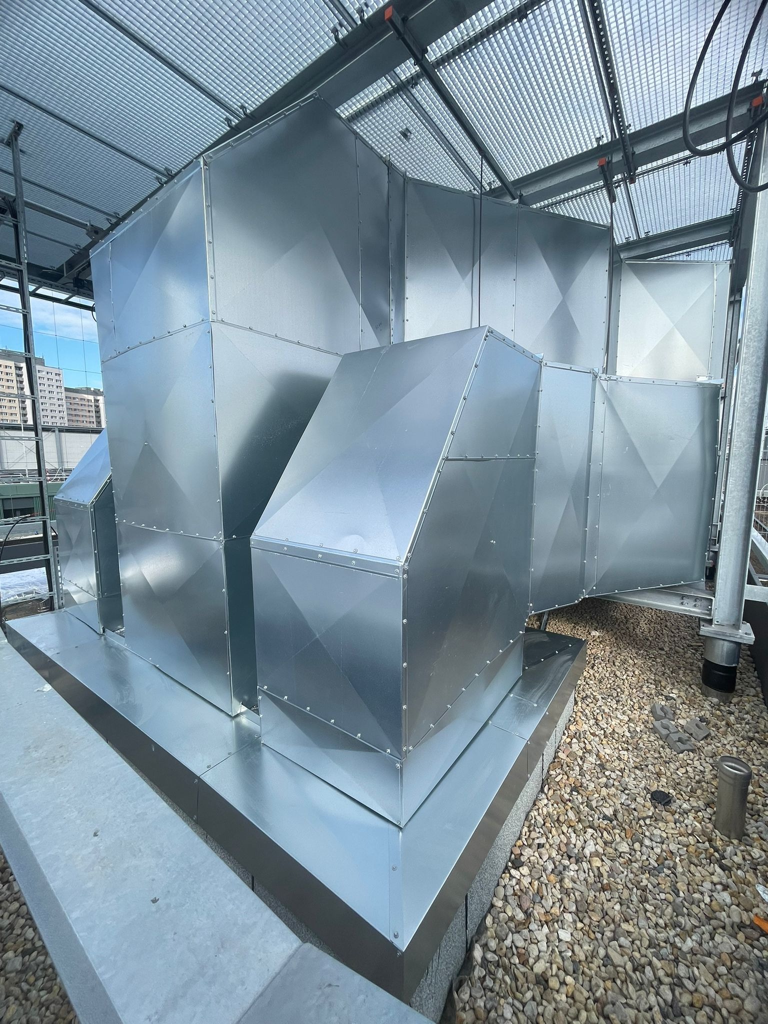 Srebrne, metalowe kanały wentylacyjne na dachu budynku, z widoczną konstrukcją wsporczą i żwirowym podłożem. Widok z dołu, w tle budynki miejskie.