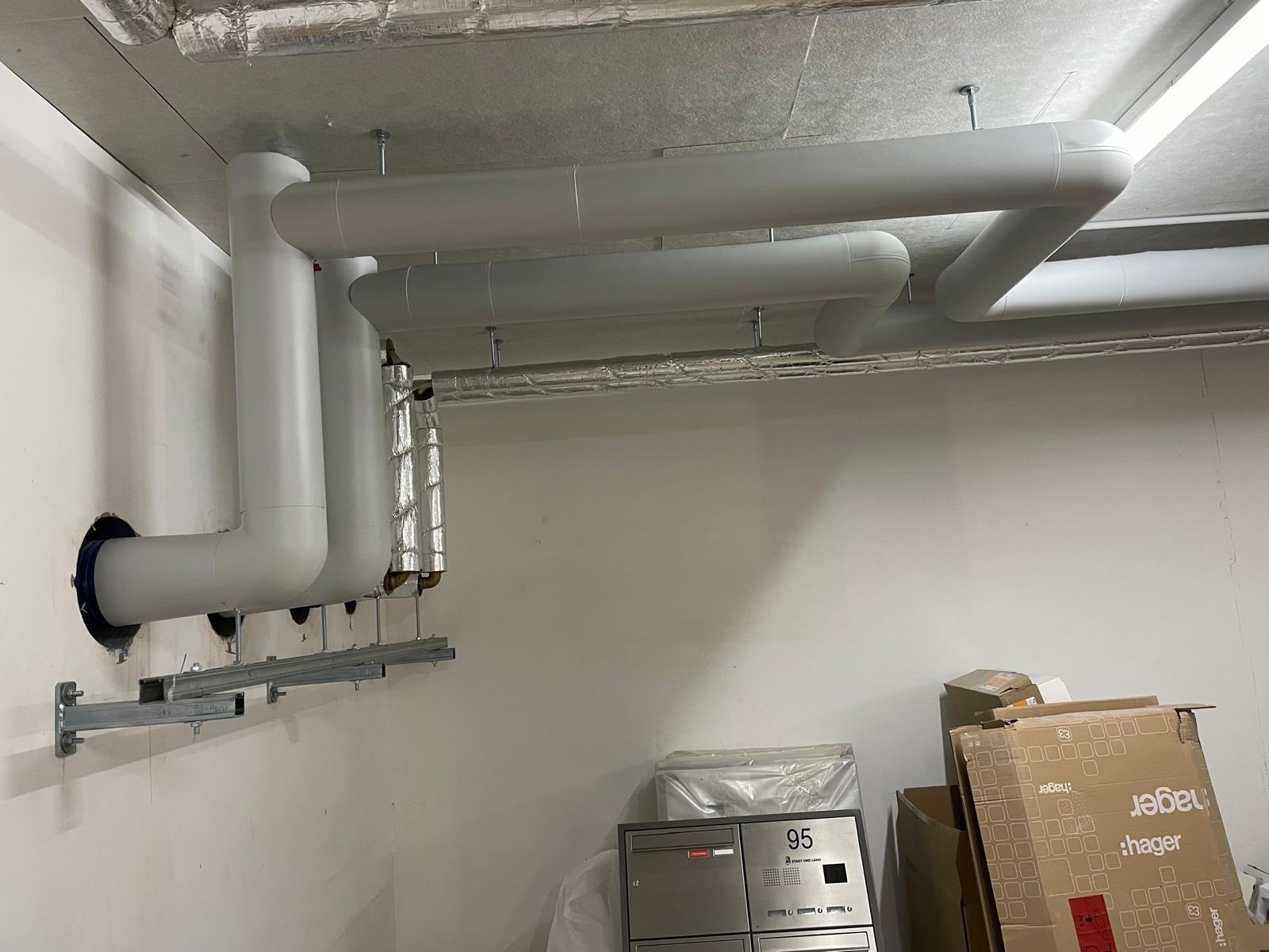 Szare rury wentylacyjne zamontowane pod sufitem w nowym budynku, widoczne skrzynki pocztowe i kartony. Instalacja w surowym stanie, przed wykończeniem.