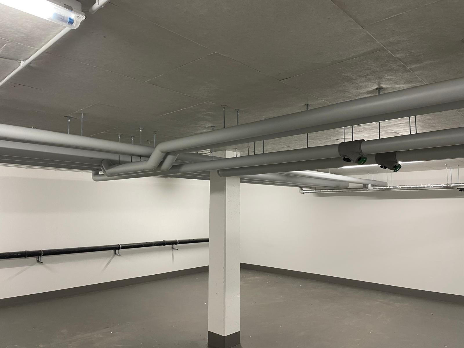 Szare rury wentylacyjne pod sufitem w nowym budynku, widoczne mocowania i oświetlenie. Białe ściany i szara podłoga. Minimalistyczne wykończenie wnętrza.