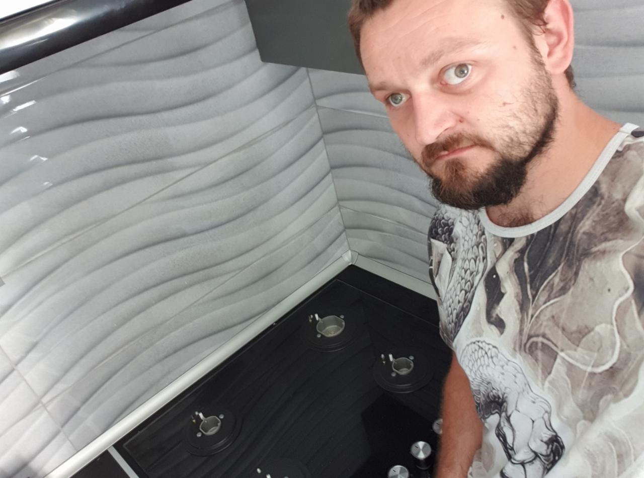 Czarna płyta gazowa z elementami palników, ułożona na tle ściany z szarymi, falistymi płytkami. Ujęcie z góry, widoczna osoba w tle.