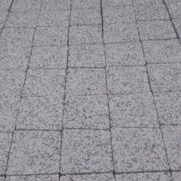Równy rząd szarych kostek brukowych o porowatej fakturze, ułożonych jedna obok drugiej pod lekkim kątem, tworzących jednolitą powierzchnię.