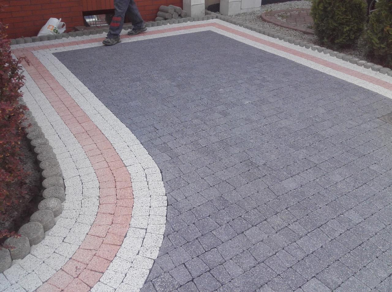 Ułożona kostka brukowa w kolorze szarym z obramowaniem z kostki białej i czerwonej, tworząca wzór na podjeździe, widoczne fragmenty osoby układającej kostkę.