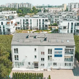 Widok z góry na budynek biurowy z szarym dachem i logo firmy na fasadzie, otoczony zielenią i zabudową mieszkalną.