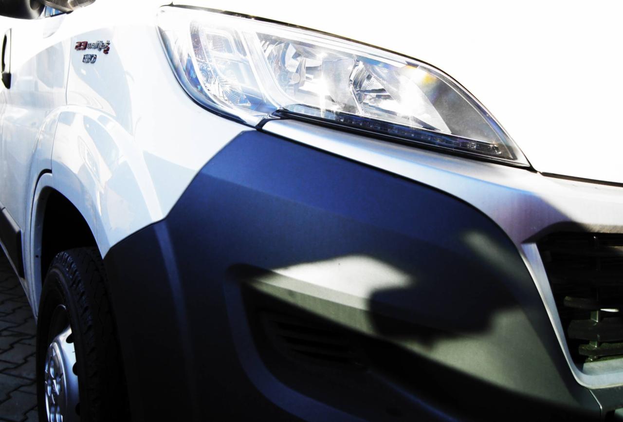 Fragment białego samochodu dostawczego z widocznym reflektorem i czarnym zderzakiem, z logo '2.3 MultiJet 130' na błotniku.