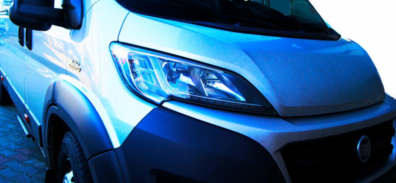 Przód białego samochodu dostawczego Fiat Ducato z widocznym reflektorem i logo na grillu, sfotografowany pod kątem.