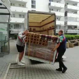 Dwóch mężczyzn w trakcie wnoszenia dużego, zabezpieczonego folią i pasami transportowymi mebla do wnętrza ciężarówki transportowej na tle nowoczesnego budynku mieszkalnego.