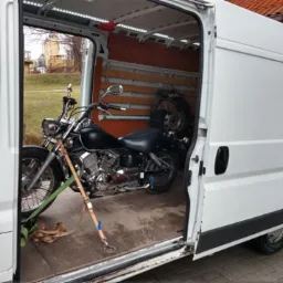 Czarny motocykl typu chopper zabezpieczony pasami wewnątrz białego furgonetki z otwartymi drzwiami przesuwnymi, widoczne również opony samochodowe w tle.