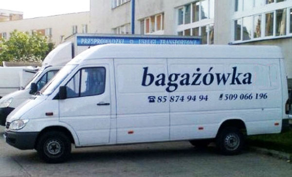 Biały bus dostawczy z napisem 'bagażówka' i numerami telefonów na boku, zaparkowany przed budynkiem.