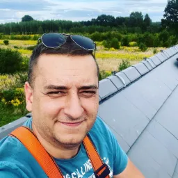 Uśmiechnięty mężczyzna w uprzęży na szarym dachu z widokiem na zielone pola i las, okulary przeciwsłoneczne na głowie.