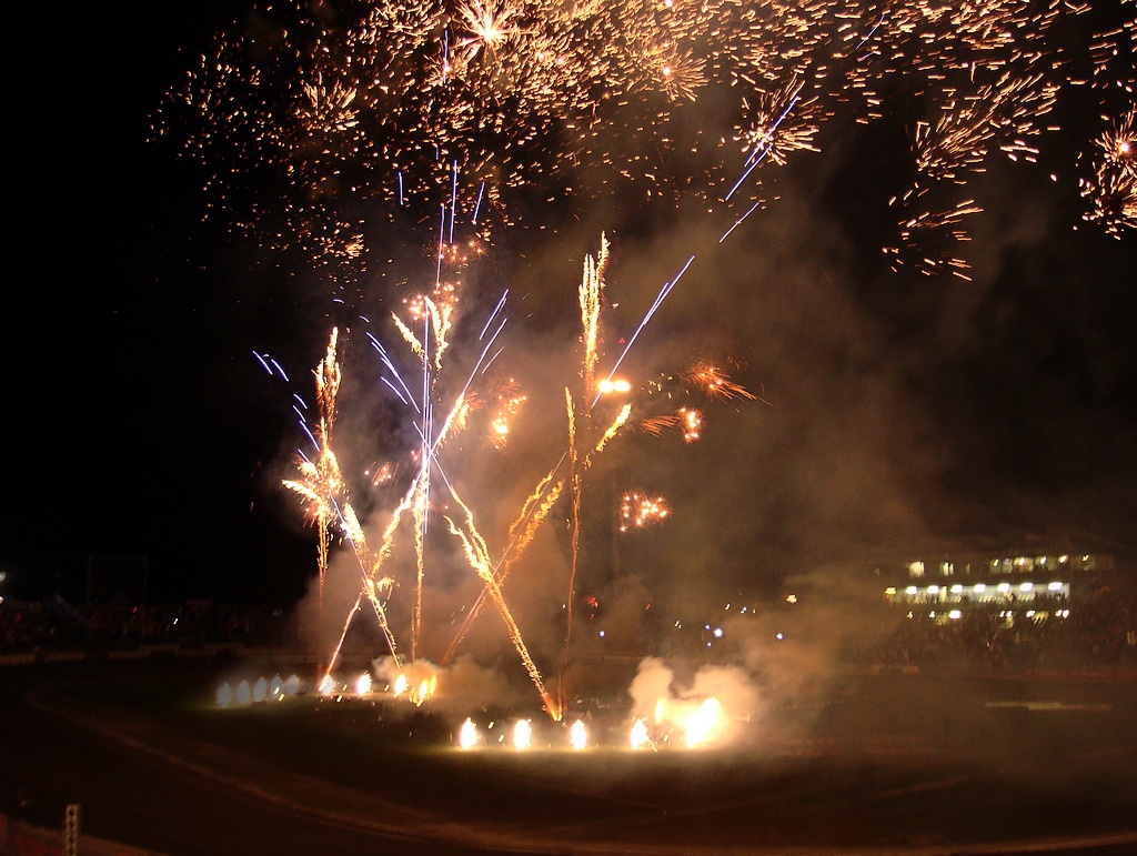 Nocny pokaz pirotechniczny z złotymi i niebieskimi fajerwerkami rozświetlającymi niebo nad torem wyścigowym, z widocznymi trybunami i budynkiem w tle.