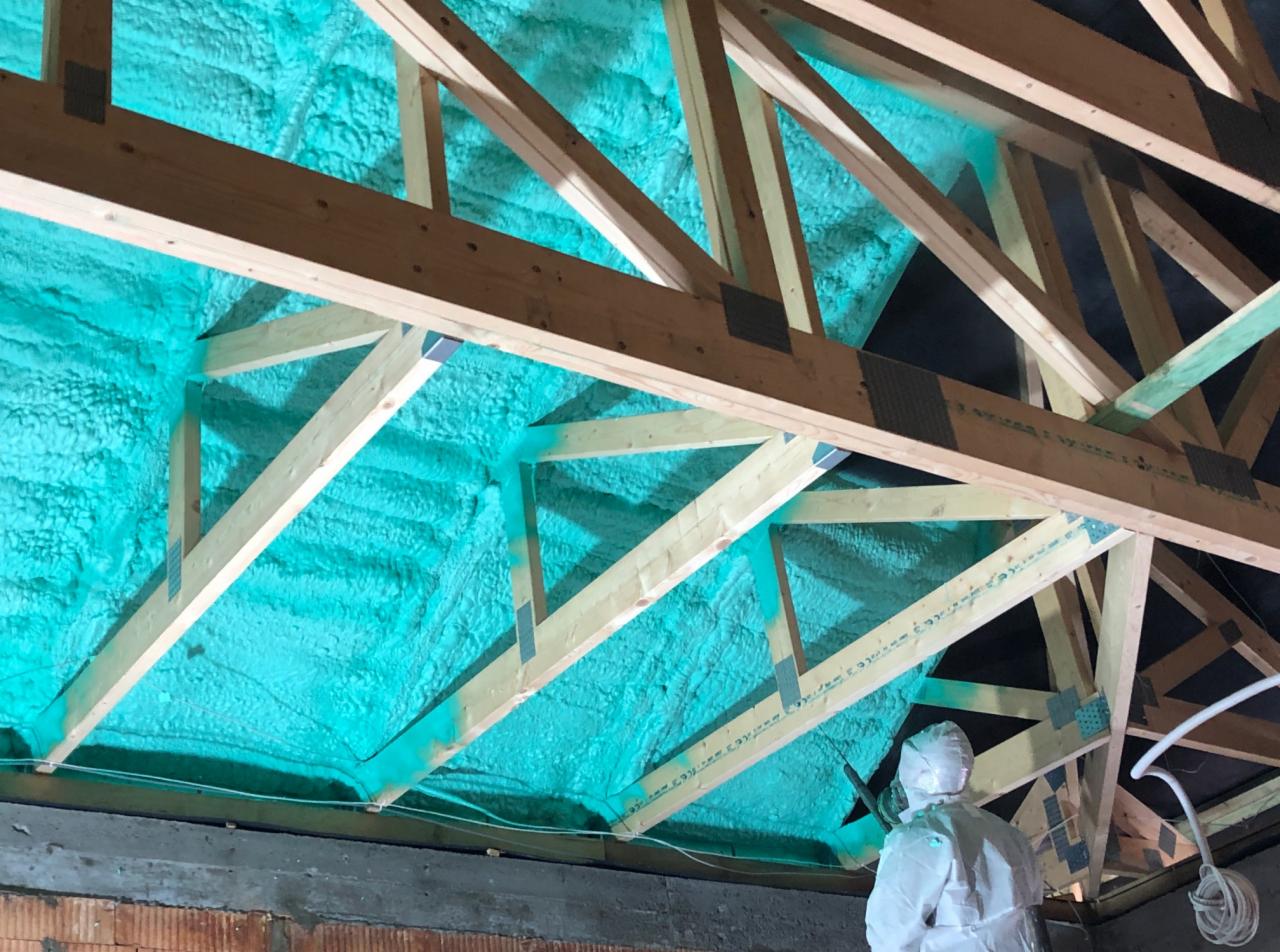 Krokwie dachowe pokryte warstwą izolacyjną, widoczny pracownik w kombinezonie ochronnym, aplikujący materiał.