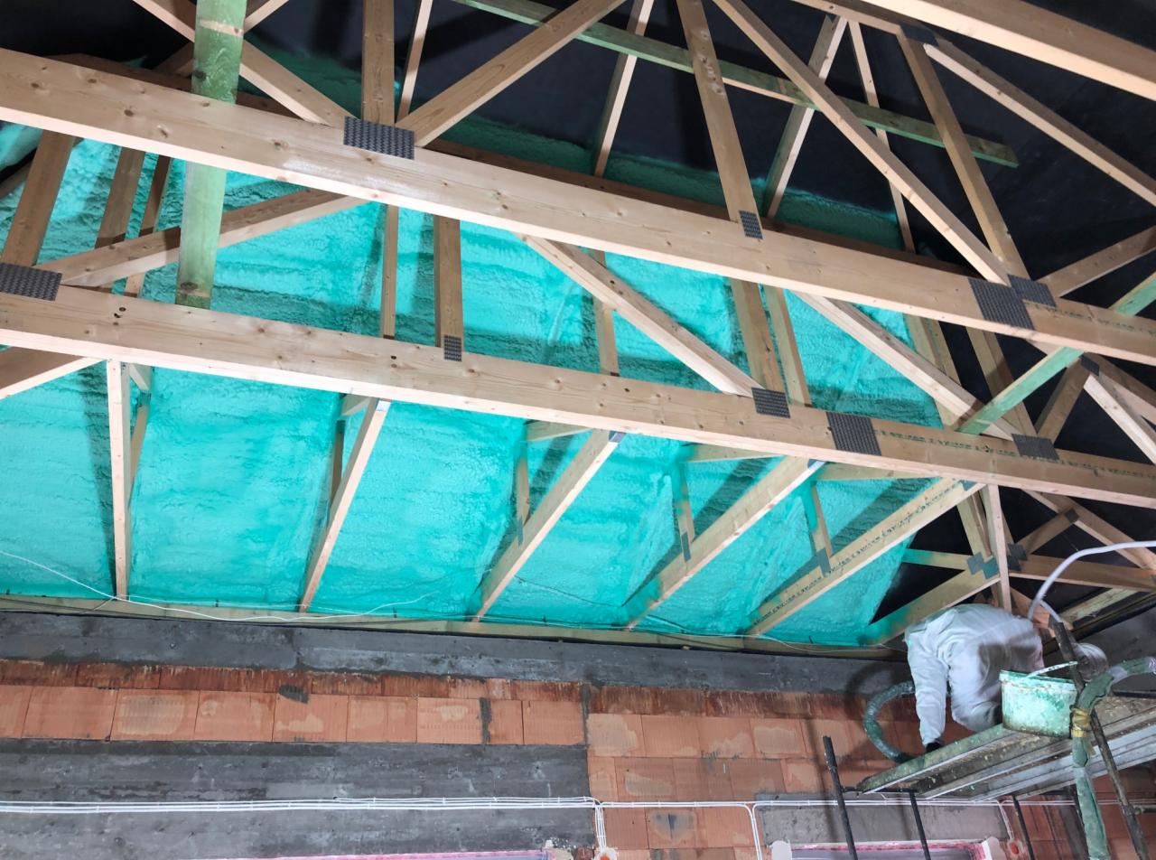 Aplikacja termoizolacyjnej, jasnozielonej piany na poddaszu, widoczna drewniana konstrukcja dachu i pracownik w kombinezonie ochronnym na rusztowaniu.