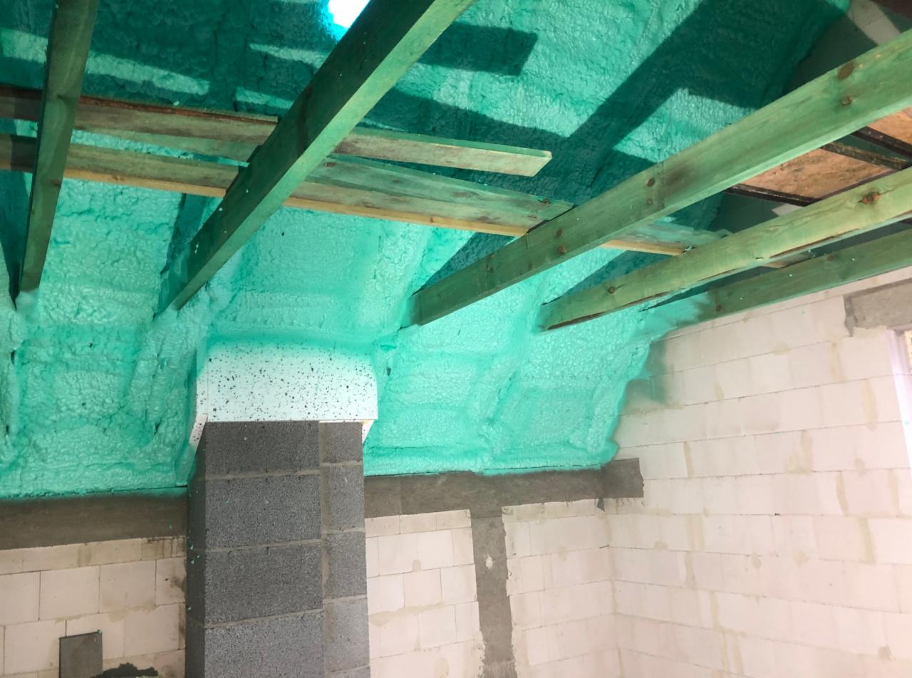 Świeżo nałożona, jasnozielona izolacja piankowa na poddaszu, widoczne drewniane krokwie i fragment komina z pustaków.