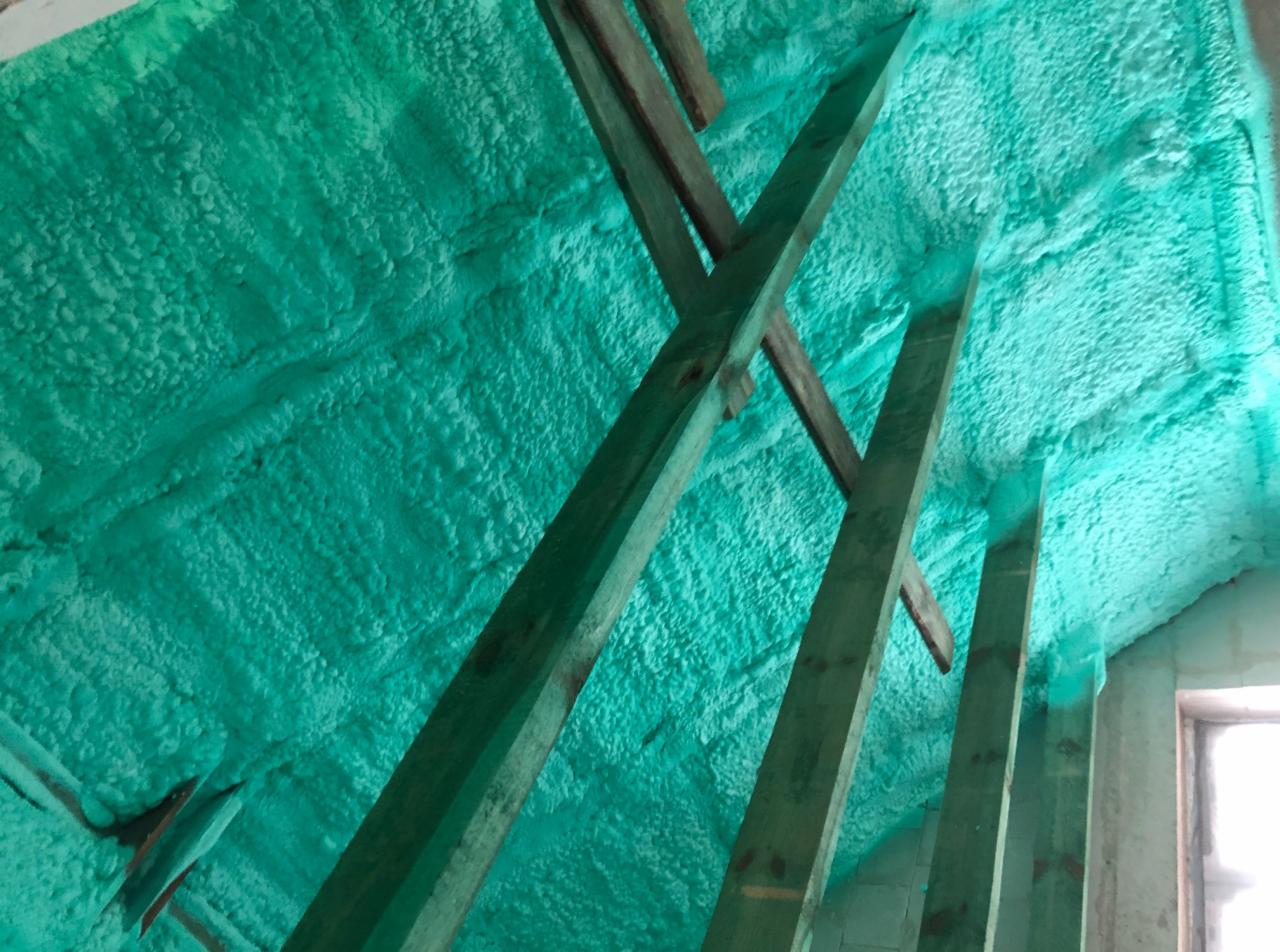 Świeżo nałożona, turkusowa izolacja natryskowa na poddaszu, widoczne elementy drewnianej konstrukcji dachu.