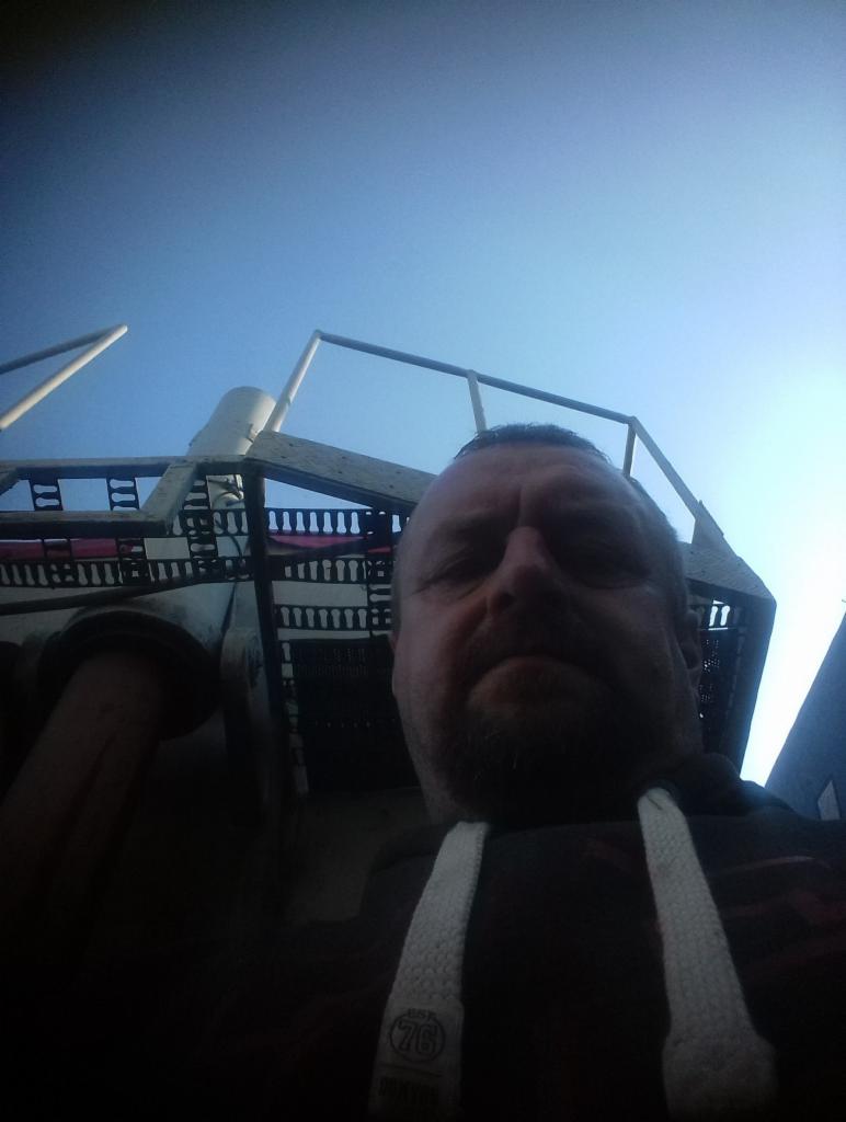 Mężczyzna robiący selfie na tle konstrukcji przypominającej statek lub rusztowanie, z widocznym niebem w tle.