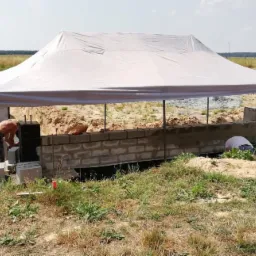 Budowa fundamentów z bloczków betonowych pod zadaszeniem na terenie budowy domu, widoczne wykopane rowy i przygotowane materiały.