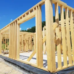 Surowa konstrukcja domu z drewnianych belek na betonowym fundamencie, widok z zewnątrz w słoneczny dzień.