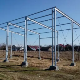 Szkielet hali stalowej na betonowych fundamentach, widok konstrukcji w surowym stanie na tle wiejskich zabudowań i błękitnego nieba.