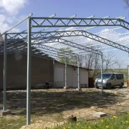 Niedokończona konstrukcja stalowa z kratownicową ramą, z betonowymi fundamentami, na tle budynków gospodarczych i zaparkowanego busa. W tle widoczna osoba siedząca na ziemi.